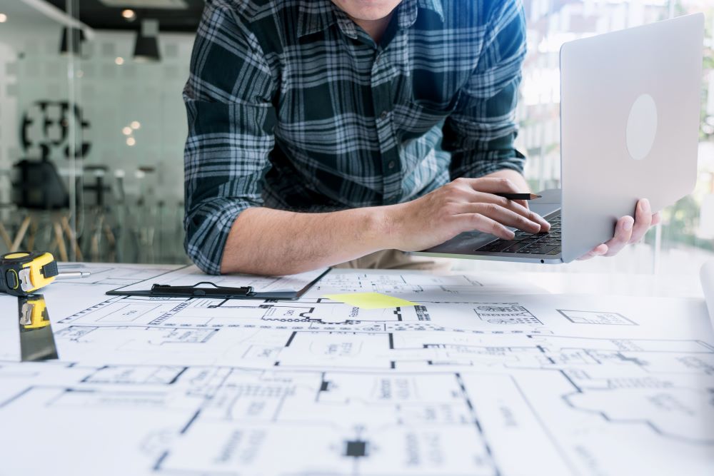 Profissional de engenharia analisando projetos em laptop ao lado de plantas técnicas, preparando o BIM Execution Plan para coordenação de projetos BIM.