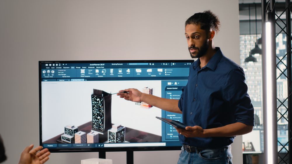 Modelador BIM apresenta modelo 3D de edifício em tela, apontando detalhes do projeto durante reunião.